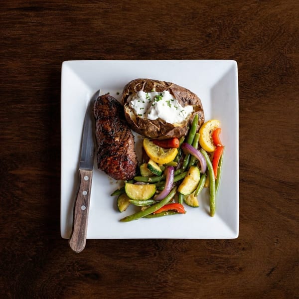 Grilled steak, baked potato with sour cream, and roasted vegetables on a white plate.