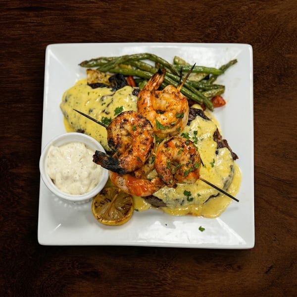 Grilled shrimp skewers, steak with yellow sauce, green beans, and a lemon slice on a white plate.