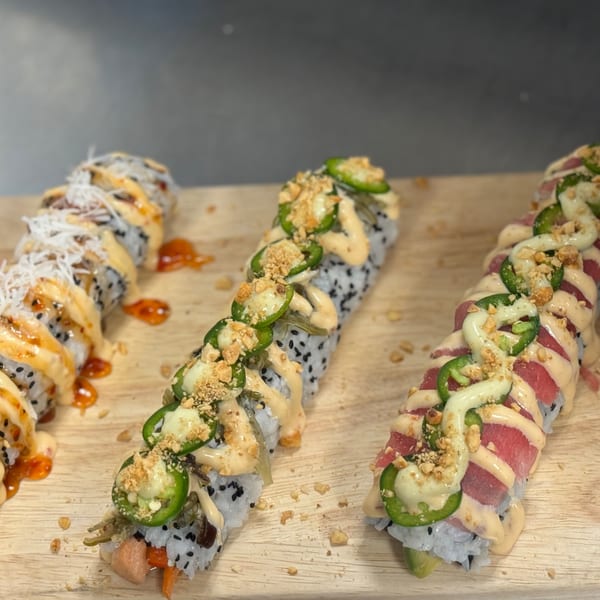 Three sushi rolls with various toppings, including jalapeños, sauces, and crunchy bits, on a wooden board.