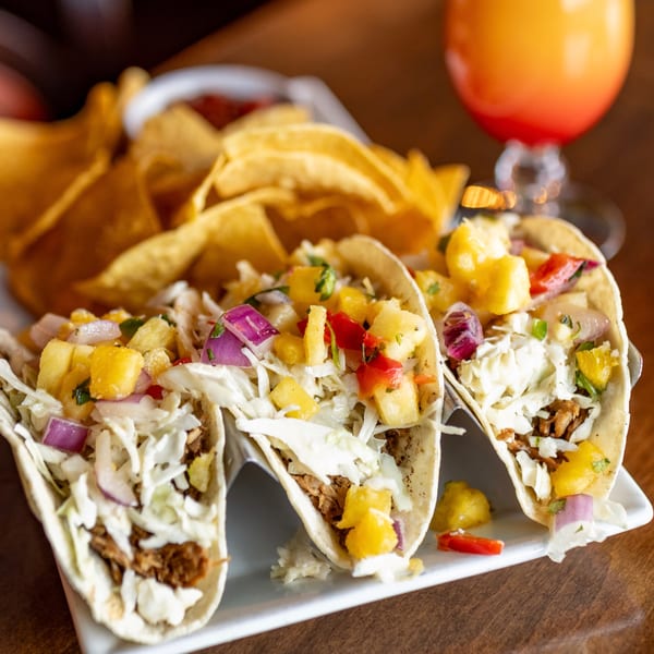 Three tacos with pulled pork, cabbage, pineapple salsa, and red onion, served with tortilla chips and a drink.
