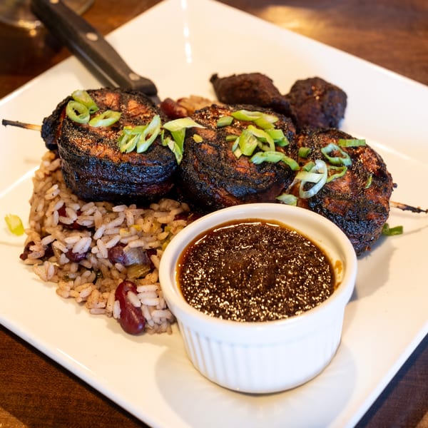 Grilled skewers of dark meat, rice and beans, and a small dish of dark sauce on a white plate.