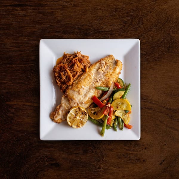 Crispy fish fillet with pulled pork, grilled lemon, and roasted vegetables on a white plate.